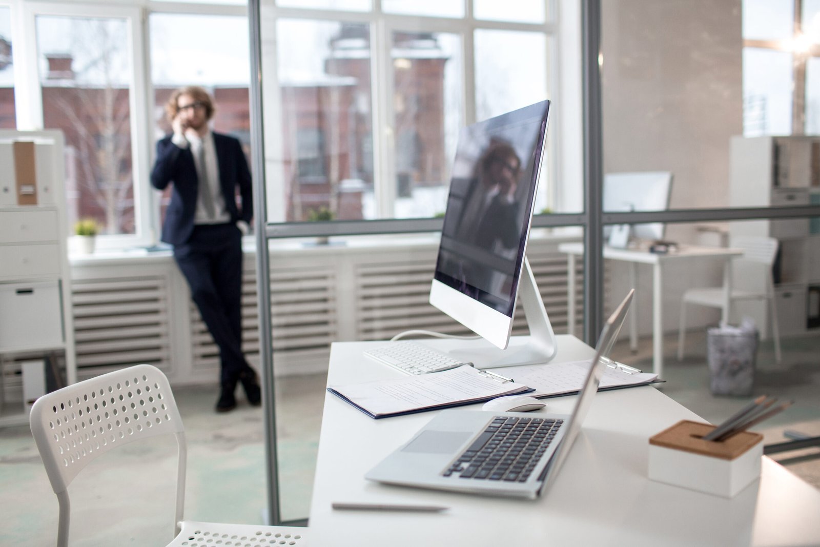 Desk of office worker with personal computer and laptop and businessman standing by window on background